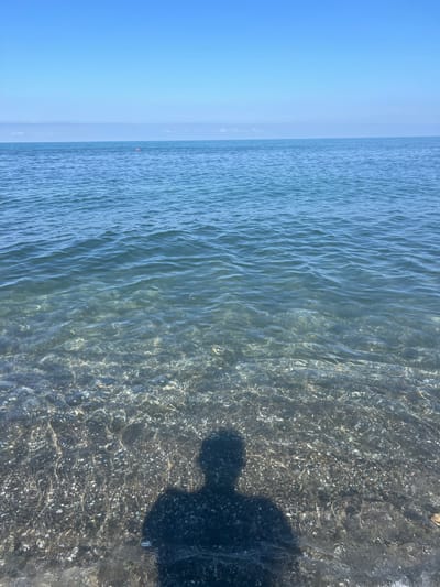 The image shows a view of the sea from the shore at Shekvetili, Georgia. The water is clear and shallow, revealing the pebbles and shells on the seabed. Gentle waves ripple across the surface, reflecting the bright blue sky. In the distance, the horizon is marked by a faint line of clouds. A single orange buoy is visible in the water. A dark shadow of a person, presumably the photographer, is cast on the shallow seabed, indicating a sunny day. The overall scene is peaceful and tranquil, characteristic of a beach setting.