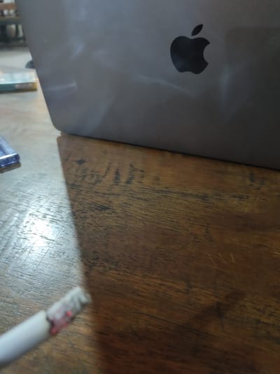 The image shows a close-up of a wooden table with an Apple laptop and a cigarette. The laptop is dark gray with the Apple logo visible. The table surface has a prominent wood grain pattern. In the foreground, the end of a white cigarette with red ash is visible, suggesting it has been recently smoked or is being smoked. A blue object, possibly a pen or stylus, is also partially visible on the left side of the frame. The lighting suggests an indoor setting, possibly in the daytime, with shadows cast by the laptop onto the table. There is no visible text in the image. The scene appears to be a casual or work-related setting.