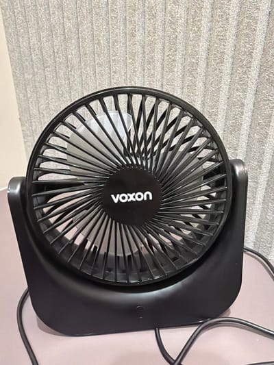 A black Vaxon brand portable fan is positioned centrally on a light pink surface. The fan has a black grille with many spokes radiating from the center. The Vaxon logo is visible in white text in the center of the grille. Behind the fan, a textured grey vertical blind is visible, suggesting the fan is indoors. The fan is connected to a black power cord which extends out of frame. The overall scene is static and appears to be an indoor setting, likely a domestic or office environment. The location in the city of Leeds, United Kingdom, is not discernible from the provided image.