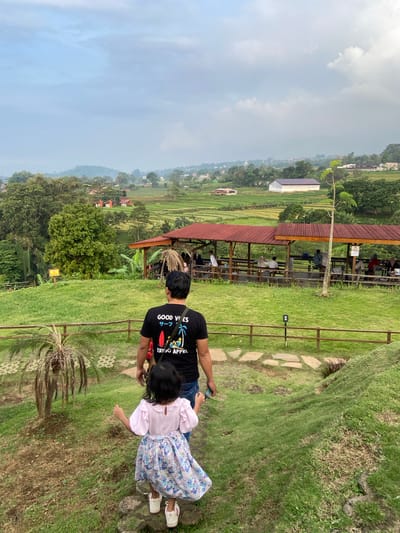 The image captures a man and a young girl standing on a grassy hillside, overlooking a landscape. The scene is located in Tamiajeng, Indonesia. The man is wearing a black t-shirt with the words "GOOD VIBES" and "ERIGO APPRL" printed on it. The girl is wearing a pink top and a dress with a floral pattern. In the background, there is a wooden structure with people seated inside. Beyond the structure, there are tiered rice paddies and houses, stretching into the distance. The sky is overcast with clouds.