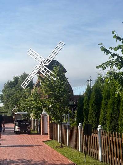 The image depicts a golf cart with the Cyrillic lettering "ГАРМОНА" on its rear parked on a red brick pathway. To the left of the cart is a wooden fence and a line of tall, green evergreen trees. In the background, a building resembling a windmill with wooden siding and white, cross-shaped blades is visible. The sky is blue with some light clouds. The location is identified as the city of Seltso, Russia.