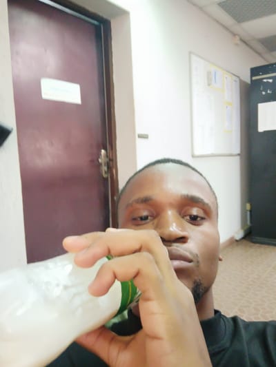 A young Black man is captured in a selfie-style shot, seen from the chest up, in an indoor setting likely within an office or institutional building in Makera, Nigeria. He is in the act of drinking from a white bottle filled with a milky white liquid, possibly yogurt or a similar beverage, which he holds to his mouth with his right hand. His left hand is also visible, possibly supporting the bottle or holding the camera. He looks directly at the lens with a neutral, possibly pensive, expression. He has short dark hair, a light mustache, and a faint goatee, and is wearing a dark, possibly black, long-sleeved top with a ribbed collar. His forehead appears somewhat shiny, possibly from lighting or perspiration.

The background shows a utilitarian interior. To the man's left is a closed dark brown door featuring a metallic handle and a rectangular white paper sign. The text on the sign is blurry and difficult to read definitively, but it appears to be a two-word phrase, possibly "PRIVATE OFFICE." Directly behind him and to his right, a white wall holds a whiteboard or bulletin board adorned with several papers, some highlighted with yellow borders and one with a blue one. Further to the right, a dark, tall piece of furniture, possibly a cabinet or partition, is visible. The ceiling above consists of light-colored square acoustic panels, and the floor is covered with a patterned, light brown or beige tile or linoleum. The overall lighting is consistent with an indoor environment, making the specific time of day indeterminate.