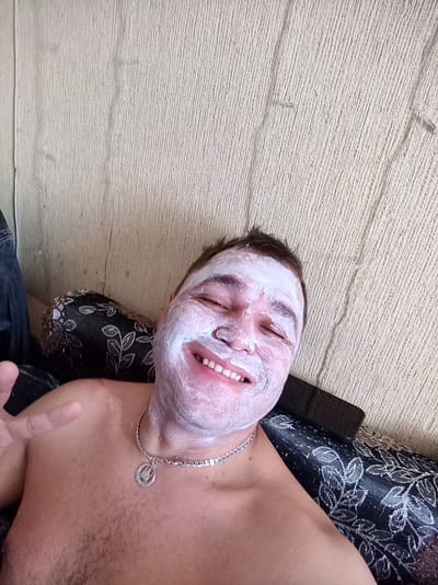 A close-up selfie captures a man with a white face mask applied to his face, smiling and looking at the camera. He is shirtless, wearing a silver chain with a pendant. He is positioned on what appears to be a couch with a patterned fabric. The background consists of a textured, light-colored wall, possibly a wallpaper or painted surface. The lighting suggests an indoor setting. There is no visible indication of the city of Beloretsk, Russia, in the image.