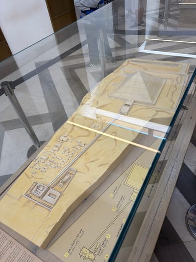 The image shows a museum display featuring a detailed architectural model of the Giza pyramid complex, specifically focusing on the pyramid of Khafre. The model is situated under a glass enclosure, with a wooden frame visible around the base. The Giza complex is located in the city of Kafrat Nassar, Egypt.

The model depicts the main pyramid, Khafre's pyramid, along with its associated mortuary temple, valley temple, and causeway. Surrounding these structures are smaller models representing ancillary pyramids and mastaba tombs, arranged in a sandy, desert-like terrain. The model is accompanied by informational diagrams or labels, some of which are partially visible and appear to contain text in both Arabic and English, identifying different components such as the "Mortuary temple," "Valley temple," and "Great Sphinx."

In the background, out of focus, are reflections on the glass enclosure, showing people standing and observing the display. The flooring in the reflection appears to be tiled with a geometric pattern of dark and light stripes. The overall environment suggests an indoor museum setting. The lighting appears to be artificial, with strip lights integrated into the display enclosure illuminating the model.