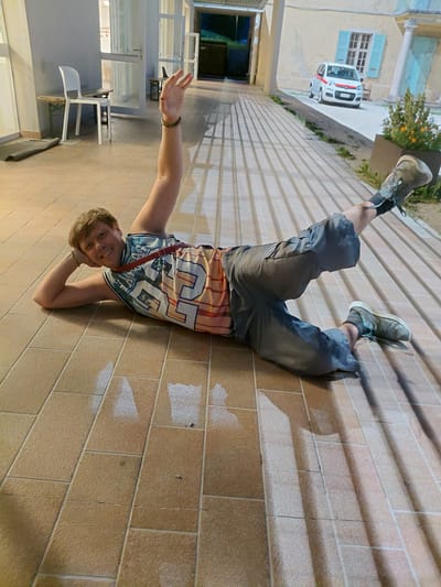 A young man is lying on his side on a tiled patio in front of a building. He is wearing a colorful basketball jersey and grey shorts. His right arm is bent and supports his head, while his left arm is raised with his hand open. His legs are bent, with his right leg extended behind him. A white car with red trim is parked in the background, with a building and some plants to the right. The scene appears to be at dusk or night, with artificial lighting casting shadows on the patio. The location is likely Levico Terme, Italy, suggested by the presence of a building with architectural features and the specific style of the car, which is a Fiat Panda often seen in Italy.