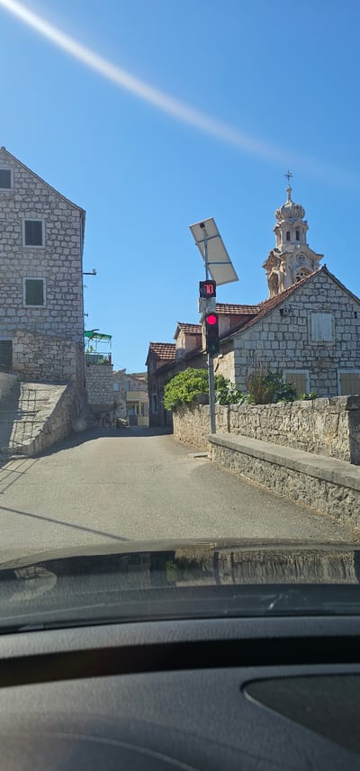 The image shows a street scene in Ložišća, Croatia. A traffic light with a red signal is illuminated, displaying the number "70" above it. The traffic light is powered by a solar panel. On the left side of the street, there is a stone building with green shutters. On the right side, another stone building with white shutters and a bell tower is visible. The street is paved and appears to be uphill. The sky is clear and blue, with a faint white streak of a cloud or contrail. The perspective is from inside a vehicle, as indicated by the reflection on the dashboard in the foreground. The architecture suggests a Mediterranean setting.