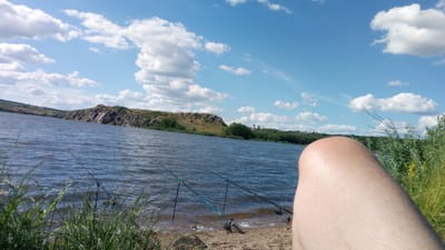 This media file captures a serene outdoor scene, likely in Buzke, Ukraine, on a bright, sunny day. The foreground is dominated by the upper part of a person's leg, with its skin displaying a healthy tan, suggesting it's a warm day. The camera angle is from the perspective of this individual, who appears to be fishing.

The mid-ground features a calm, blue body of water, possibly a river or lake, rippling gently under the sky. Several fishing rods are set up along the shoreline, their tips pointing towards the water, indicating an ongoing fishing activity. The shoreline itself is composed of pebbles and sand, with lush green grass and other vegetation growing in abundance, framing the scene from the bottom left and right.

In the background, a rocky hill rises from the opposite bank of the water. The hill is covered in dry grass and scattered trees, adding to the natural beauty of the landscape. The sky above is a vibrant blue, dotted with fluffy white clouds, characteristic of good weather. The overall impression is one of peace and relaxation, a typical scene for a leisurely day spent fishing outdoors. The time of day appears to be late morning or early afternoon, judging by the bright sunlight and clear sky. There is no visible text in the image.