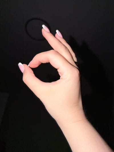 A person's left hand, with French manicured nails, forms an "OK" gesture against a dark background. A faint circular shadow is visible in the upper left quadrant. The scene is not specific to the city of Razumnoye, Russia.