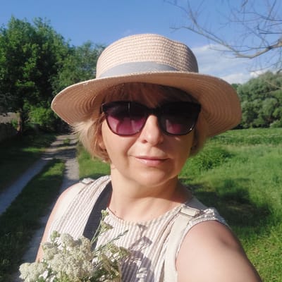 This is a slightly rotated, medium shot photo taken outdoors in a park or natural setting in Voronkiv, Ukraine. The main subject is a woman, viewed from the chest up, wearing a straw hat and sunglasses. She has blonde hair, and her lips are slightly parted. She is wearing a striped, light-colored top with thin straps. In her right hand, she is holding a small bouquet of delicate white wildflowers.

The background is lush and green, with grass and trees. The sky is a bright blue, indicating it is daytime with clear weather. A path, possibly made of gravel or stone, winds through the green area. The overall impression is of a pleasant, sunny day spent outdoors. There is no visible text in the image.