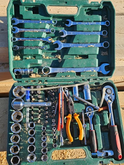 An open, green plastic toolbox filled with various metal tools, including wrenches, sockets, pliers, and a retractable knife, rests on a surface of light-colored wooden planks. Sawdust is scattered throughout the toolbox and on the surrounding wooden surface. The toolbox is positioned outdoors, suggested by the natural lighting and the wooden planks which appear to be construction material or firewood. The scene does not provide any specific information to link it to the city of Mayya, Russia, or any particular urban or rural setting. There are no people or actions depicted in the image.