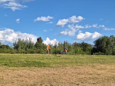 This image depicts a sunny day in Markkleeberg, Germany, likely in a park or natural area. Two individuals are cycling on a grassy embankment. The person on the left is wearing an orange shirt and dark shorts, cycling a bicycle with no visible trailer. The person on the right is wearing a dark t-shirt and orange shorts, cycling a bicycle with a small black trailer attached. They are positioned on a slight incline with a line of trees and bushes forming the background. The foreground consists of dry, yellowed grass, transitioning to greener grass further up the embankment. The sky above is a clear blue with scattered white clouds.