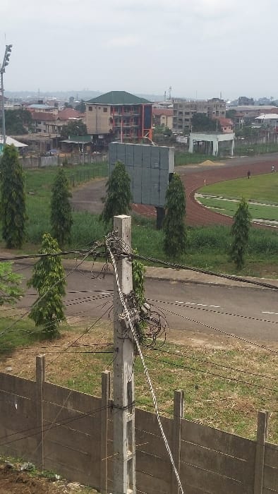 The image shows a view of a concrete utility pole with multiple wires running from it. The pole is located in front of a concrete wall, with a grassy area in the foreground. There is a dirt road to the left of the pole, and a track and field stadium to the right. The stadium has a red track and a green field. The field has several trees on it, and there is a building in the distance. The image was taken in the city of Buea, Cameroon, at the coordinates 4.163 latitude and 9.279 longitude. There is one figure in the image, a person walking on the track and field stadium. The time of day is daytime and the weather appears to be cloudy. There are no other people in the image.