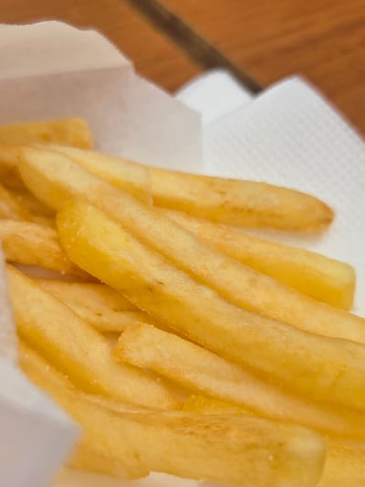 A close-up, eye-level shot shows a white paper container filled with golden french fries. The fries are arranged in a slightly overlapping manner, with some appearing to be dusted with salt. The container's white paper has a slightly textured surface. In the background, a wooden surface is visible, suggesting a table or countertop, and its warm brown tones are slightly out of focus. The overall lighting appears to be natural, casting soft shadows and highlighting the crispy texture of the fries. This image was taken in Sergiyev Posad, Russia.