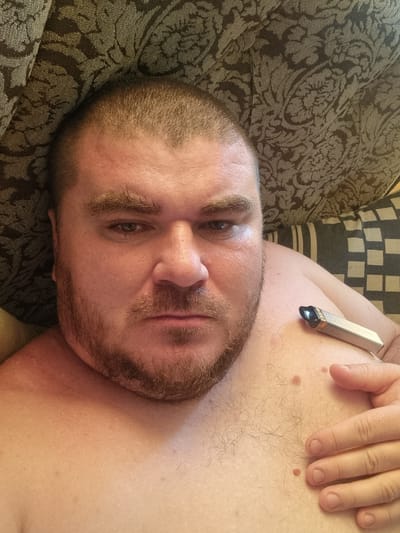 The image shows a close-up, overhead selfie of a man lying down, shirtless. He has a shaved head, light brown hair on his face, and visible chest hair. He is holding a silver electronic cigarette device to his chest with his right hand. The background consists of a patterned sofa. The image is not identifiable as being taken in Romny, Ukraine.