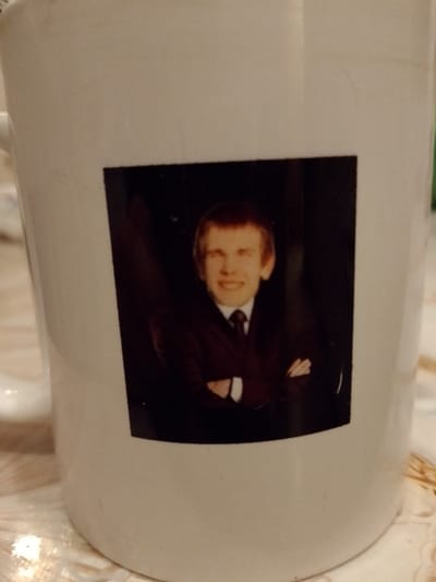 The image displays a white ceramic mug with a photograph printed on its side. The mug has a slight sheen and shows some minor smudges or marks above the photo, suggesting it might be in use or handled frequently.
The printed photograph features a young man from the chest up, against a dark, possibly black, background. He appears to have short, light brown or blondish hair styled with bangs swept forward. The man is smiling broadly, showing his teeth, and his eyes are light-colored, though the exact color is not clearly discernible due to resolution and lighting. He is wearing formal attire: a dark suit jacket over a white shirt and a dark tie. His arms are crossed over his chest, and he is looking directly at the camera with a cheerful and confident expression.
The mug is placed on a surface that appears to be a light-colored, possibly patterned tablecloth or countertop, featuring muted tones of beige, white, and light brown, suggesting an indoor setting. The overall lighting seems to be artificial, illuminating the mug and the surface it rests upon.