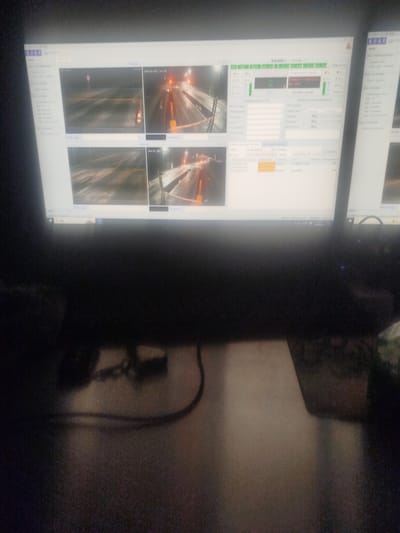 A computer monitor displays a multi-camera surveillance system. The screen is divided into four quadrants, each showing a nighttime view of a road. The road surfaces appear wet or icy, and there are orange traffic cones and a pink circular marker visible in some frames. The monitor's interface shows various controls and text, some of which is in a non-English script. The monitor is set up on a desk, with cables and other objects blurred in the foreground. The image is dimly lit, suggesting the scene is in a dark environment. The specific location of Түндүк айланып өтүүчү жол 84 га, Kyrgyzstan is not discernible from the visual information in the image.