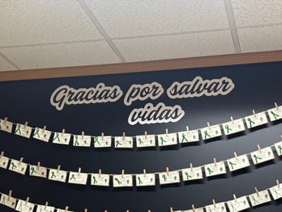 A dark blue wall displays the silver lettering "Gracias por salvar vidas." Below it, several rows of small cards hang on strings, secured with miniature wooden clothespins. Each card features a green ribbon, some bearing visible names like "Estela," "Juliann," and "Carlos Miguel." The cards seem to honor or acknowledge individuals, with the ribbons possibly signifying a cause or awareness. The setting is an indoor space with a white, textured ceiling panel.