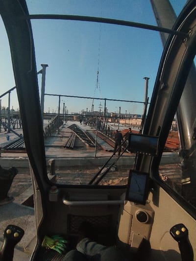 The image depicts an active industrial construction site in Navoiy City, Uzbekistan, viewed from the interior of a heavy equipment cabin. The foreground shows parts of the cabin's interior, including a control joystick on the left, a digital display panel on the right, and the operator's legs clad in blue jeans, with a green work glove resting on the floor.

Outside the cabin, under a clear blue sky, a large-scale construction area is visible. The site features extensive concrete foundations and numerous erected steel structural columns, some appearing as I-beams and others cylindrical. Stacks of pre-fabricated metal trusses and beams are laid out on these concrete platforms, extending into the midground. A crane hook, suspended by a cable, is visible overhead, indicating active lifting operations.

Multiple individuals, identified as construction workers in dark clothing, are present on the concrete platforms among the building materials. At least three workers are discernible; one worker is standing near a pile of metal beams, while others are further down the platform. In the distant background, industrial structures, utility poles, and power lines are visible, with some faint traces of smoke or exhaust rising. The ground around the concrete structures consists of reddish-brown earth.