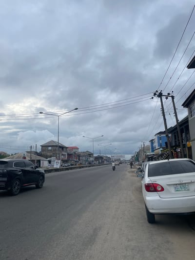 This image captures a street scene in Rumuepirikom, Nigeria, under a cloudy sky. A two-lane asphalt road runs through the center, with a sidewalk on the right. Several vehicles are visible, including a black SUV on the left side of the road, a white Toyota sedan parked on the right, and other cars and a motorcycle further down the road. Buildings line both sides of the street, featuring a mix of architectural styles, some appearing commercial with signs and awnings. Utility poles with multiple wires are prominent along the roadside. Pedestrians are present on the sidewalk. The overall atmosphere is that of a busy urban environment.