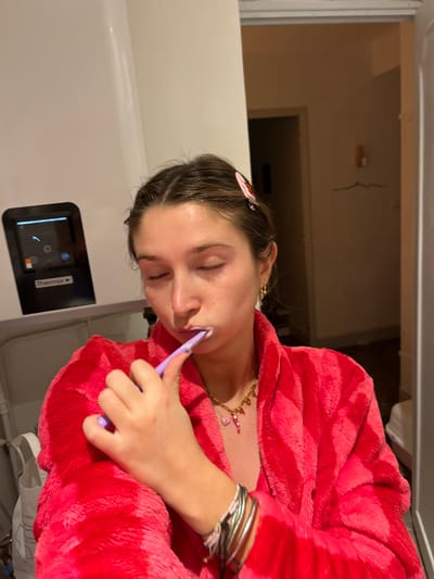 A young woman in a red bathrobe brushes her teeth with her eyes closed. She has a strawberry clip in her hair, gold chain necklace with charms, and bracelets. The woman is next to a white cabinet with a digital display panel, possibly a water heater. A doorway is visible in the background.