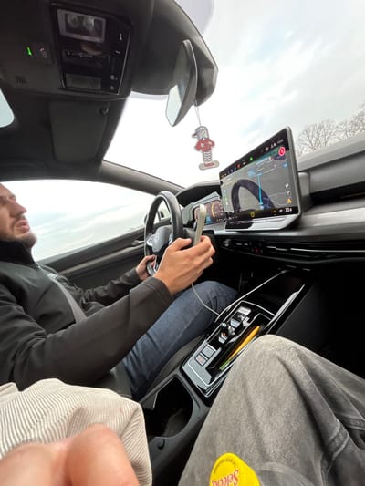 The image depicts the interior of a modern vehicle from the perspective of the front passenger seat. The location is La Wantzenau, France, as indicated on the vehicle's vertically oriented infotainment screen, which displays a navigation map with a blue route line. The time displayed on the screen is 15:26, and the external temperature is 22.0 degrees.
A male driver is visible on the left, wearing a dark, long-sleeved, collared jacket and blue jeans. He is secured by a seatbelt. His left hand is positioned on the multi-function steering wheel, which bears a Volkswagen emblem. His right hand holds a light-colored smartphone, connected by a white cable that extends downwards. The driver's head is turned slightly to the right, and he has a beard or stubble.
The front passenger's lower torso and legs are partially visible on the right, clad in light grey trousers. A yellow container with the partial label "Selecto" is resting on the passenger's lap.
The vehicle's interior features a digital instrument cluster behind the steering wheel, a sleek black center console with illuminated controls, and a small yellow object (possibly a pen) lodged in a console slot. A character-shaped air freshener, depicting a figure with a red cap on a spring, hangs from the rearview mirror. An overhead console is visible, showing a green illuminated indicator next to what appears to be an SOS button. The sky visible through the windshield is overcast.