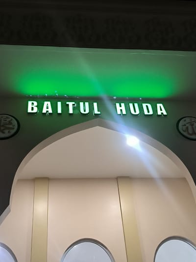 The image captures an architectural interior, likely an entrance or hallway, characterized by a prominent archway. Above the arch, a sign illuminated with a vibrant green light displays the words "BAITUL HUDA" in bold, raised letters. The green light casts a luminous glow on the wall behind the sign.
On either side of the archway, circular decorative plaques are affixed to the wall. The plaque on the left features the name "Muhammad" written in elegant Arabic calligraphy, while the plaque on the right displays the word "Allah" in similar Arabic script. The archway itself is constructed from light beige material, with a vertical light-colored column or panel framing its base on the left side. A bright lens flare, originating from an unseen light source, is visible to the right of the arch's highest point. The ceiling above the sign is dark and appears to have a subtle textured pattern.
No people or specific activities are depicted. The artificial lighting, including the glowing green sign and the lens flare, suggests the image was taken indoors or during the evening/night. Given the text "Baitul Huda" (meaning "House of Guidance" in Arabic) and the presence of Islamic calligraphy, the location is strongly indicative of a mosque or an Islamic center in Mee Tanjong Usi, Indonesia.