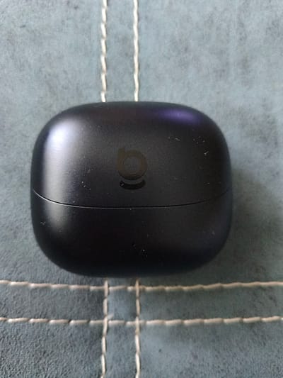 A close-up, overhead shot shows a black charging case for wireless earbuds resting on a textured, grey surface with white stitching. The charging case is oval-shaped with a prominent seam running horizontally across the middle, indicating it is likely open or about to open. A subtle, dark logo resembling a stylized 'b' or a rounded infinity symbol is visible on the upper half of the case. The surface beneath the case appears to be a fabric or mat with a grid pattern formed by the white stitching. The lighting suggests it is an indoor setting with soft illumination, creating gentle reflections on the curved surface of the charging case. The image provides no indication of the city of Otradny, Russia, or any environmental context beyond the immediate surface.