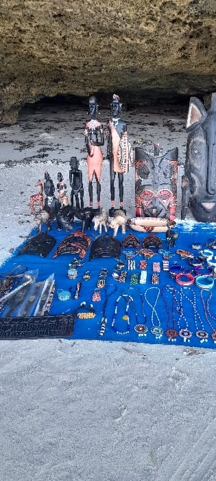 The scene, located in Ziwa La Ng'ombe ward, Kenya, depicts an outdoor display of artisanal crafts on a sandy beach. The foreground is composed of light grey sand. Across the midground, a blue fabric is spread out, acting as a display base for numerous items. These items include a variety of beaded necklaces, bracelets, and small decorative objects, some individually packaged in clear plastic sleeves. Immediately behind the blue fabric, arranged on what appears to be a slightly raised surface or the sand, is an assortment of larger carved wooden artifacts. This collection features two tall, slender wooden figures resembling Maasai warriors, depicted with traditional attire, shields, and spears. Alongside these are several smaller carved wooden animal figures, including giraffes and elephants, and various smaller human figurines. To the right, multiple large, dark carved wooden masks with intricate details, along with other decorative carved wooden panels and bowls, are displayed. The background shows a sandy area with scattered dark debris, leading to a prominent light-colored rocky cliff face or overhang that contains a shadowed recess or cave opening. No individuals are visible within the frame.
