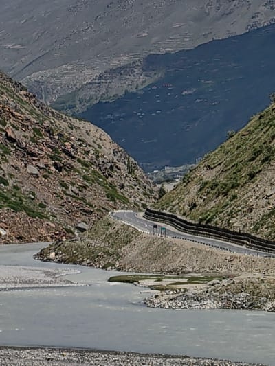 The image depicts a scenic mountain landscape with a winding road and a river. In the foreground, a wide, light-colored river flows, with a rocky shore on the left side and a sandy bank on the right. The river appears to be shallow and fast-flowing, with visible ripples on its surface.
A paved road, bordered by a guardrail and a stone wall, curves along the side of a steep, rocky mountain on the right. The road is well-maintained and shows signs of traffic, with at least one vehicle and a person visible. The mountain slopes are covered in sparse vegetation, with patches of green grass and small shrubs.
In the background, more mountains rise, their slopes terraced and dotted with small settlements. The overall impression is one of a remote and rugged natural environment, potentially in a mountainous region of India, such as Burwa, known for its dramatic topography and winding roads. The lighting suggests it is daytime, with sunlight casting shadows and highlighting the textures of the rocks and vegetation.