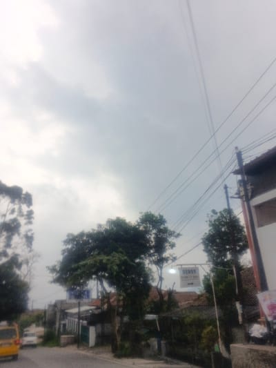 This image depicts an outdoor urban or semi-urban scene under an overcast sky in the city of Morrumbala, Mozambique. The dominant visual element is a light grey, uniformly cloudy sky occupying the upper half of the frame.

A paved road is visible along the lower left and center, with a blurry yellow vehicle (possibly a van) partially visible on the far left, followed by a lighter-colored car further back. On the right side, the edge of a multi-story building with white and darker sections is prominent, featuring utility poles and multiple overhead power lines or communication cables crisscrossing the upper right quadrant against the sky.

In the mid-ground, numerous trees with dense green foliage are scattered throughout, contributing to a lush environment. A rectangular, illuminated sign hangs suspended over a lower structure, appearing to be a commercial or service establishment. The text on the sign is partially legible, with "BIDAN" visible at the top, followed by smaller, indistinct text (possibly "HOME CARE") and an arrow pointing to the right. Buildings with reddish-brown tiled roofs are visible behind the trees and the sign, receding into the background. The overall impression is one of overcast daylight with no discernible individuals or specific actions taking place.
