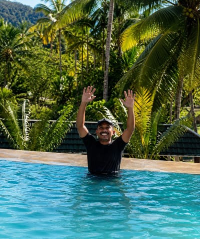 A man is standing waist-deep in a bright blue swimming pool, with his arms raised in the air and his hands open. He is wearing a black t-shirt and a dark baseball cap. The man is smiling broadly at the camera. Behind him, lush green palm trees and dense tropical foliage fill the background, with hints of mountains in the distance under a clear blue sky. The poolside is visible in the foreground, a light brown, textured surface. The scene appears to be in a tropical resort setting, possibly in Silaiyajae, Indonesia, given the abundant tropical vegetation.