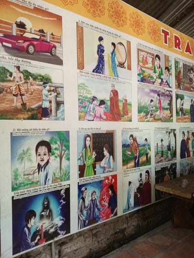 The image is a photograph of a large, colorful mural displayed on a wall. The mural is divided into multiple panels, each illustrating a scene with accompanying text in Vietnamese and English. The top of the mural features decorative floral patterns and the letters "TRA" in large red lettering, suggesting it is part of a larger display or project.
The mural appears to depict moral or karmic lessons, with each panel illustrating a cause and effect scenario related to past lives and current circumstances. Some of the depicted scenes include:
* A red convertible car with ships in the background, labeled "What about transportation?"
* Two women looking at their reflections in a mirror, with the text "What cause is possessing opulent clothes in this life?"
* People working on what looks like a road, with text about roads for people in previous lives.
* A group of people offering robes to monks, with text about offering robes to Buddhist monks and nuns in previous lives.
* A family offering food at a table, with text about generously giving aims to the poor in previous lives.
* A child with a harelip, with text "What is the cause of being harelipped?"
* A person blowing out a Buddhist candle, with text about blowing out a Buddhist candle with bad-smelled mouth in previous lives.
* Two people scolding and cursing at each other, with text about scolding and cursing their parents in previous lives.
The overall style of the mural is illustrative, with simplified figures and bright colors. The image is taken from a slightly angled, eye-level perspective, looking upwards at the mural. The background shows a textured wall and what appears to be part of a wooden structure above the mural, suggesting it is displayed indoors or in a covered outdoor area. The lower part of the image shows a tiled floor and a wooden plank or shelf. The lighting is somewhat dim, casting soft shadows.
The mural is located in Nam Ban, Vietnam, as indicated by the location information provided.