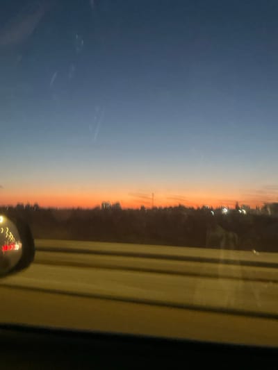 The image depicts a dusk urban scene from the perspective of a moving vehicle, looking towards the city of Hod Hasharon, Israel. The sky displays a gradient from deep blue at the top, transitioning through lighter blue to a prominent band of orange and red along the horizon, indicative of sunset. Faint, elongated streaks and light glints are visible across the upper portion of the sky, consistent with reflections or imperfections on a window. Along the horizon, a dark, blurred silhouette of buildings and trees defines the urban outline of Hod Hasharon. Scattered within this silhouette are several small, bright, blurred artificial lights. The lower foreground features a significantly blurred, light-colored surface with parallel dark lines, representing a multi-lane road or highway, conveying a sense of rapid forward motion. On the extreme left, a portion of a vehicle's side-view mirror is visible, reflecting indistinct red and white lights.