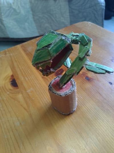A handmade, green, cardboard carnivorous plant model is positioned on a light brown, wooden table. The plant has a distinct head with sharp, jagged teeth and a red interior, supported by a segmented green stem. The stem is rooted in a cylindrical brown cardboard base with a red interior visible at the top. The background consists of a blurred, light-colored sofa and a portion of a wall. The image is captured from a high angle, with the plant in the foreground. There are no people present in the image. The location is Nicorești, Romania.