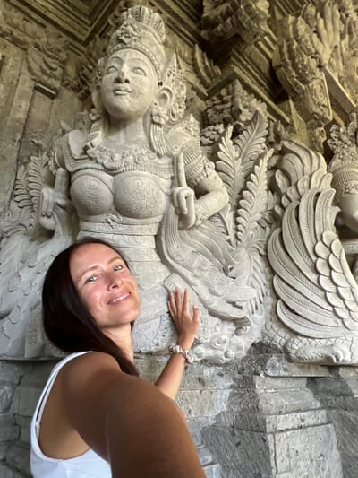 A woman takes a selfie with a bas-relief sculpture in Kedewatan, Indonesia. The sculpture features a female figure with ornate details, including elaborate headdress, chest ornamentation, and flowing garments, and she reclines amidst foliage and winged figures. The surrounding architecture consists of carved stone elements. The woman, with dark hair and a white tank top, smiles at the camera.