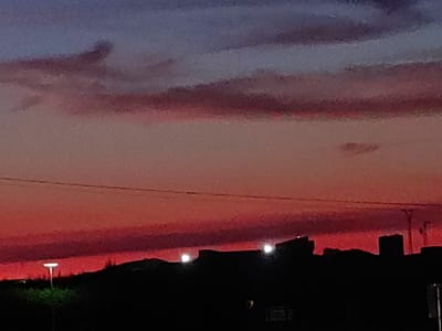 The image depicts a dramatic sunset over Valverde de Leganés, Spain. The sky transitions from a deep indigo at the top to vibrant reds and oranges near the horizon, with layered clouds streaking across the sky. The silhouette of buildings forms the lower edge of the image, with two bright lights visible, possibly from streetlights or illuminated windows. A single streetlight is also visible in the foreground, casting a glow on the dark landscape below. The scene is characterized by its strong contrasts between light and shadow, typical of twilight.