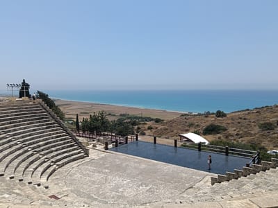 A person stands near a reflective pool in an ancient amphitheater with a view of the sea. The amphitheater's stone seating is prominently featured in the foreground, curving around an open performance area. The pool, a dark, rectangular surface, reflects the sky and surrounding structures, bordered by a railing. Beyond the amphitheater, a coastal landscape unfolds. Dry, rolling hills covered in scrub and scattered trees lead down to a stretch of cultivated land and then the blue expanse of the Mediterranean Sea. The sky is clear and a pale blue, suggesting a bright, sunny day, likely midday due to the strong light and shadows. The overall mood is serene and historical, showcasing a blend of ancient architecture and natural beauty. No visible text is present.