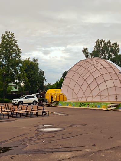 The image depicts an outdoor event space in Khotkovo, Russia. In the foreground, rows of empty wooden benches with metal frames are arranged on a paved area. Further back, a white SUV with its trunk open is parked. To the right of the SUV is a large, translucent dome structure with a geodesic frame, suggesting it might be a temporary pavilion or stage cover. Adjacent to the dome is a yellow, tent-like structure. A person is seated on one of the benches facing away from the camera, and another person stands near the yellow structure. The background features mature trees and a building, and the sky is overcast with clouds. The ground in front of the benches shows several puddles. A decorative banner with text and cartoon-like illustrations is visible along the front of the stage area.