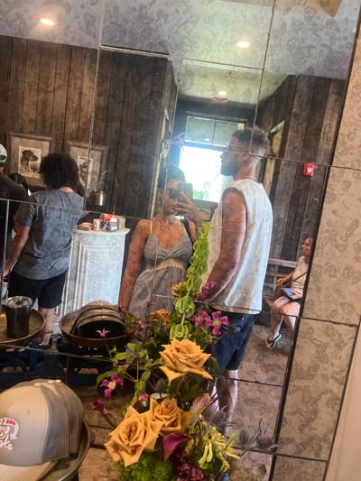 A medium shot captures a reflection of a couple standing in what appears to be a rustic-themed establishment. The man, on the right, is wearing a sleeveless white t-shirt and dark shorts, while the woman, on the left, is in a striped dress and sunglasses. She is holding a phone, suggesting she is taking the photo. In the background, visible through the mirror, is a wooden-paneled wall adorned with framed pictures, a decorative fireplace, and a lamp. A woman is seated in the background, observing. In the foreground, a bouquet of flowers, including roses and clematis, sits on a table, along with a hat and a cup. The location is identified as Palm Valley, United States of America.