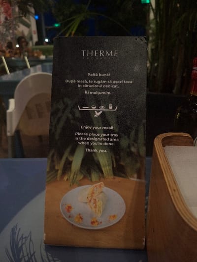 The image depicts a close-up, eye-level view of a dining area, identified as Therme București, located within or adjacent to Săftica, Romania. A dark, rectangular standing sign, exhibiting minor edge wear, is centrally positioned. The sign displays "THERME BUCURESTI" at the top, followed by instructions in Romanian and English: "Enjoy your meal! Please place your tray in the designated area when you're done. Thank you." An icon depicting a tray, plate, drink, and cutlery points to a disposal cart. The lower portion of the sign features a photograph of a cut wrap or burrito on a white plate, garnished with orange and yellow components, set against a wooden surface.

The sign rests on a blue-grey table surface, which has a subtle light-colored pattern on its left side. To the right of the sign, a light-colored wooden napkin holder containing white napkins is partially visible. A dark glass bottle with a red label is discernible in the blurred background to the right. The overall background is out of focus, revealing a dimly lit environment, likely evening or night. Green foliage, resembling large plants, occupies the mid-background. Distant, indistinct human figures are seated at tables in the far left background, indicating other patrons. Blurred white chairs and tables are also present. Diffused light sources appear as blue and green orbs in the deep background.