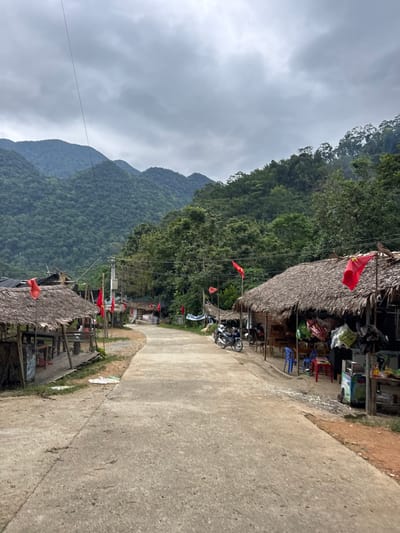 The image depicts a scene on a paved road in Cổ Lũng Commune, Vietnam, likely a rural area due to the surrounding lush green mountains. The sky is overcast with dense clouds. On both sides of the road, there are small shops with thatched roofs. Red flags with yellow stars, the national flag of Vietnam, are prominently displayed, some on poles and others attached to the structures. Several motorcycles are parked along the roadside in front of the shops. The shops appear to be selling various goods, with items visible on shelves and tables outside. The overall atmosphere suggests a quiet, perhaps slightly remote, community in a mountainous region.