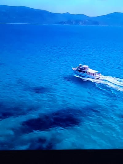 The image shows a wide shot of a bright blue sea with a white boat sailing across it, leaving a white wake behind it. In the distance, there are mountains under a clear sky. The scene is not located in Sambir, Ukraine, as Sambir is an inland city.