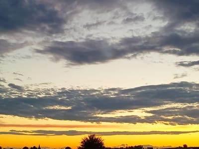 This is an outdoor photograph taken at sunset in Somaglia, Italy. The image captures a wide sky filled with dramatic clouds. A bright, intense yellow and orange glow emanates from the horizon, suggesting the sun is very low. Layered above this vibrant band are darker, greyish clouds, some of which are illuminated from beneath by the sunset's light. The upper portion of the sky transitions to a lighter, more neutral tone with wispy clouds. In the lower part of the frame, a silhouetted treeline stretches across the landscape, interrupted by the faint outlines of buildings and a prominent church spire. A single, small, dark object, possibly an airplane, is visible in the upper sky.