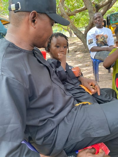 An adult male wearing a black baseball cap and a black traditional tunic is seated on the left, looking towards a young child positioned on his lap or directly beside him. The child, with braided hair, is dressed in a black garment similar to the adult's, holding a small orange cup in their left hand and placing their right hand near their mouth. The adult's right hand rests on his leg, holding a yellow tassel-like object. In the mid-ground, to the right of the primary subjects, another adult male is seated on a blue plastic stool. He wears a white t-shirt with a multi-figure graphic print and light-colored jeans. A portion of a yellow and green auto-rickshaw is visible behind him. The scene is outdoors in Ochadam, Nigeria, characterized by dappled sunlight filtering through overhead tree foliage onto a bare earth ground.