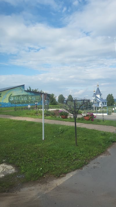 The image depicts an outdoor scene in the village of Kvarsa, Russia, featuring a mural on the side of a building, a pathway, a church, and lush green grass under a partly cloudy sky. In the foreground, a paved road runs along the right edge, with a grassy area to its left. A white pole stands in the grassy area, supporting a sign with a mural. The mural, visible on the left side of the image, is a large painting on the wall of a building depicting a ship with people on board, surrounded by a stylized blue wave and green elements. A pathway, possibly made of concrete or gravel, runs horizontally across the middle ground of the image, separating the grassy area from the background.

In the middle ground, two individuals are walking on the pathway. One person, wearing a light-colored shirt and dark pants, is closer to the mural. Another individual, dressed in dark clothing, is further down the path. Two decorative metal stands with hanging flower baskets are positioned along the path. The baskets contain blooming flowers in shades of pink and white.

In the background, on the right side of the image, a white church with a blue roof and blue domes stands prominently. A golden cross tops the central dome. The church is surrounded by evergreen trees and appears to be part of a larger religious complex or grounds, with a white fence visible in front of it. The overall environment suggests a peaceful, rural setting during the daytime, with a blend of man-made structures and natural elements.