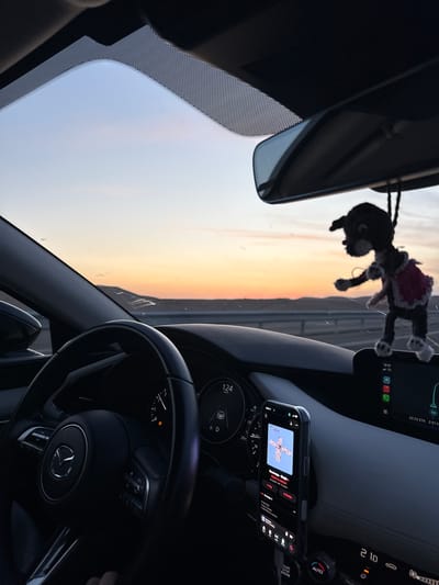 Captured from inside a car, the image features the driver's perspective of a Mazda dashboard. The dashboard includes a steering wheel, instrument panel, smartphone attached to a holder displaying a music application, and a central console. A fuzzy doll is suspended from the rearview mirror. The windshield frames an outdoor view with a sky transitioning from light purple to orange, possibly indicating sunrise or sunset. The location may be Hevel Eilot Regional Council, Israel, given the time and date on the dashboard suggest it is 19:31 and the phone displays weather information relevant to the region.