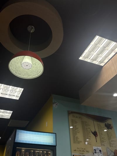 The image captures the interior ceiling and upper wall sections of "Flavors of Aventura," an ice cream and yogurt shop located at 2921 NE 207th St, Aventura, Florida, United States. The ceiling is dark, possibly black, with a large, illuminated red dome-shaped pendant light suspended from it. The interior surface of the pendant light is white with a detailed, embossed floral pattern. Above the pendant light, a decorative series of concentric circular wooden trim elements is affixed to the ceiling. Several rectangular fluorescent light panels are visible, some actively emitting light. The upper wall transitions from a yellow section on the left to a light blue/teal section on the right. A white, multi-lens security camera, appearing to resemble a smiling face, is mounted on the upper light blue wall. Below the camera, a large menu board titled "Flavors" displays various ice cream and yogurt options, along with text including "Ice Cream with a Healthy Twist." On the far left, the top of a commercial refrigerator unit with a light blue tiled upper surface and visible bottled beverages is present. No individuals or specific actions are depicted within the frame.