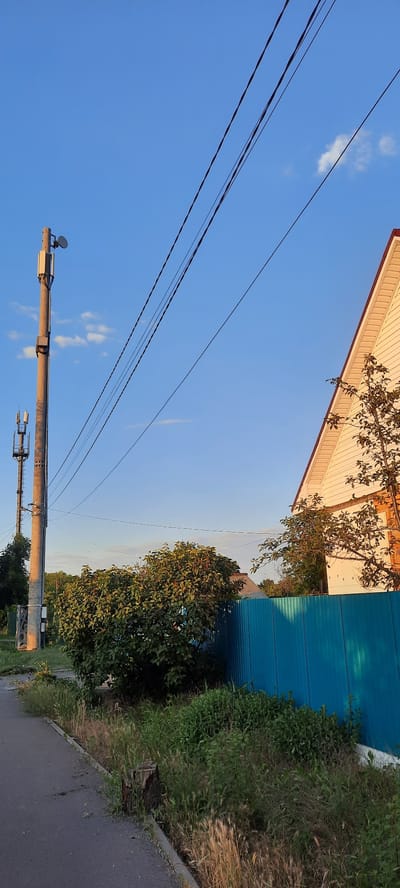 The image depicts a rural or suburban street scene under a clear blue sky with scattered white clouds. A paved road runs along the left side of the frame, bordered by overgrown grass and vegetation. A prominent wooden utility pole, equipped with cell phone antennas and dishes, stands near the road. In the background, a white house with a red-trimmed roof is visible, partially obscured by trees and a bright blue metal fence. Another utility pole, also with antennas, is visible further down the road. The scene appears to be located in Shakhty City District, Russia, given the presence of typical infrastructure and vegetation. No people are present in the image.