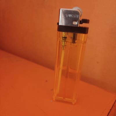 The image shows a yellow lighter on a bright orange surface. The lighter is made of plastic and metal and is transparent. The brand "TINKAI" is printed on the metal part. The surface appears to be a smooth, flat, and horizontal surface. The image was taken in an indoor setting. The location, based on the coordinates provided, is in Plumbon, Indonesia. There are no people or other objects visible in the image. The lighter is in a vertical position, and the flame is not lit. It appears that the lighter is sitting still and there is no motion or interaction in the image. The surface is clean and free of any debris or other objects.