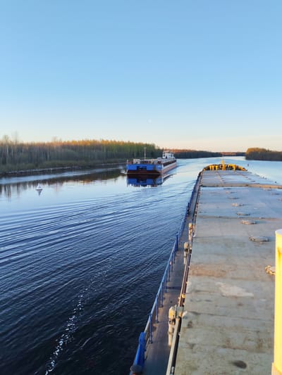 A long shot captures two barges moving along a river near Kurdiug, Russia. The barge closest to the viewer is gray and occupies a significant portion of the right side of the frame. The other barge is a darker blue and is further away, moving in the opposite direction. Both vessels leave wakes in the water. The river reflects the clear blue sky. Trees line the riverbanks, with autumn colors beginning to appear on some of the leaves. A small, white buoy is visible in the river near the left bank. The time of day appears to be late afternoon.